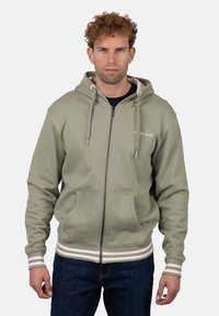 Sudadera con cremallera de color verde oliva con capucha ajustable con cordón, bolsillos tipo canguro y detalles de rayas blancas en los puños y el dobladillo. Textura de tela suave.