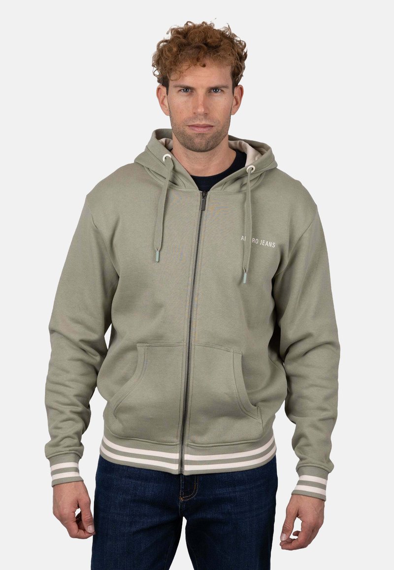 Sudadera con cremallera de color verde oliva con capucha ajustable con cordón, bolsillos tipo canguro y detalles de rayas blancas en los puños y el dobladillo. Textura de tela suave.