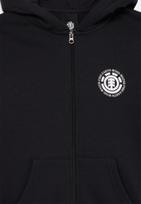 La felpa nera con zip presenta una tasca frontale e un logo circolare con motivi ambientali sul lato sinistro. Tessuto dalla texture liscia visibile.