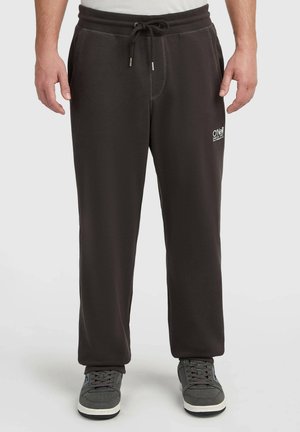 Man draagt zwarte joggingbroek met trekkoord en zijzakken en grijze sneakers, staand tegen een effen lichte achtergrond.