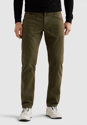 Hombre con pantalones verde oliva con grandes bolsillos delanteros y zapatillas blancas, de pie contra un fondo claro y liso.