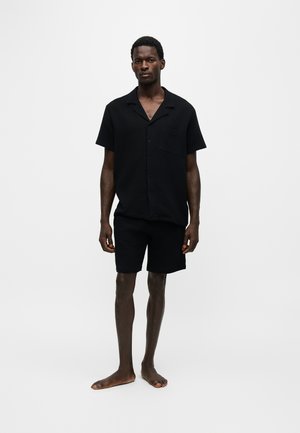 Homme debout pieds nus portant une chemise noire à manches courtes boutonnée et un short noir sur un fond blanc uni.
