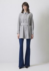 Grå långärmad skjortklänning med knapplist, bälte i midjan och en dekorativ nål. Kombinerad med utsvängda blå denimjeans och svarta klackar.