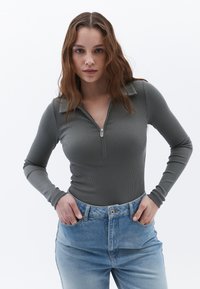 Langärmliges, geripptes Oberteil in Dunkelgrün mit einem Reißverschlusskragen. Kombiniert mit hellblauen High-Waist-Jeans mit einem Knopf und zwei Fronttaschen.