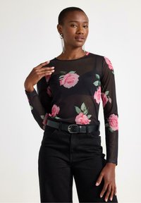 Sort langærmet mesh top med pink rosemønster, parret med sorte høj taljede jeans og et sort bælte. Model iført iøjnefaldende øreringe.