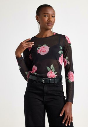 Sort langærmet mesh top med pink rosemønster, parret med sorte høj taljede jeans og et sort bælte. Model iført iøjnefaldende øreringe.