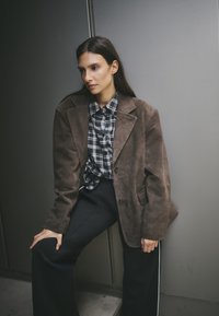 Bruine suède blazer over een zwart-wit geruite blouse, gecombineerd met zwarte wijde broek met witte zijkanten. Soepele textuur.