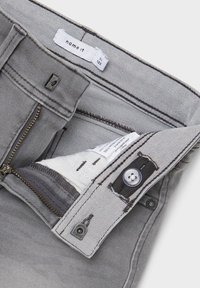 Gray denim trousers featuring a zipper with gold teeth, button closure, and contrasting stitching. Interior label shows brand and size information.