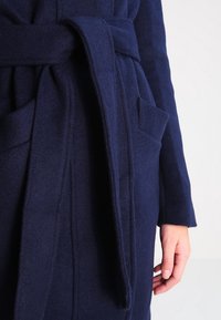 Marinblå ullkappa med knytning i midjan, sidoficka och mjuk textur med en enkel design och lös passform.