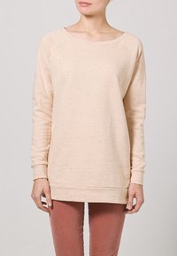 Světlé béžové tričko z měkké látky, s kulatým výstřihem a dlouhými rukávy s mírně volným střihem. Na látce jsou malé skvrnky.