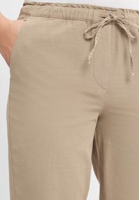 Gros plan sur un pantalon beige à cordon de serrage avec une main dans la poche gauche, montrant la texture du tissu et les détails de la ceinture élastique.