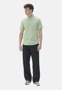 Camicia verde chiaro a maniche corte con bottoni e taschino, abbinata a pantaloni neri larghi e sneakers grigie. Design semplice e look casual.