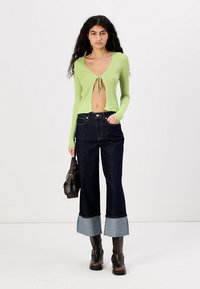 Top corto verde de canalé con lazo en el frente, combinado con jeans de mezclilla oscuros con la pierna acampanada y puños en azul claro, complementado con botas negras hasta el tobillo.
