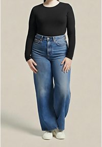 Personne portant un haut noir à manches longues, un jean bleu taille haute à jambes larges et des chaussures blanches, debout devant un fond uni.