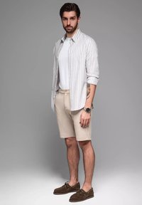 Camisa ligera de rayas abotonada en blanco y gris, combinada con pantalones cortos beige y mocasines marrón oscuro. Presenta un reloj en la muñeca.
