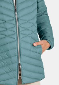 Close-up of teal quilted jacket with double zipper and hand partially in side pocket.