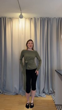 Femme debout devant des rideaux gris, portant un haut à manches longues vert olive avec un lien à la taille et un pantalon court noir, pieds nus sur un sol en bois.