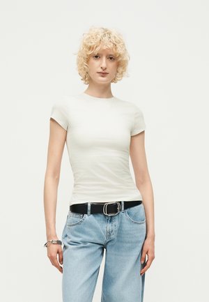 Άτομο με κοντά σγουρά ξανθά μαλλιά που φοράει εφαρμοστή λευκή κοντομάνικη μπλούζα, ανοιχτό μπλε τζιν, μαύρη ζώνη και ασημένια βραχιόλια.