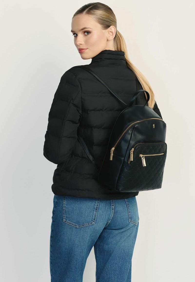 Czarny pikowany skórzany plecak z złotymi zamkami i akcentami, noszony przez osobę w czarnej kurtce puchowej i niebieskich dżinsach.