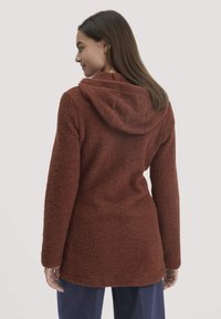 Eine Frau, die mit dem Rücken zur Kamera steht, trägt eine lange, strukturierte, rostfarbene Kapuzenjacke über einer dunkelblauen Hose vor einem schlichten, hellen Hintergrund.