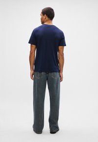 Homem de costas, vestindo uma t-shirt azul-marinho simples de manga curta e jeans azuis desgastados de corte solto, contra um fundo branco.
