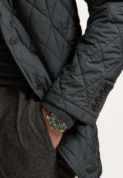 Mann in einer gepolsterten, dunklen Jacke und grauem Pullover mit der Hand in der Tasche, die eine Armbanduhr mit grünem Zifferblatt und braunem Lederarmband zeigt.