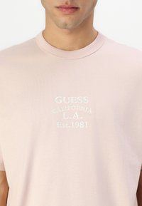 Rozā kokvilnas T-krekls ar apaļu kaklu, īsām piedurknēm un izšutu dizainu "GUESS CALIFORNIA L.A. Est. 1981" baltā krāsā.