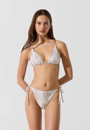 Młoda kobieta o długich brązowych włosach, ubrana w jasno szary bikini w wzór węża z przewiązanymi bokami, stojąca na jednolitym jasnym tle.
