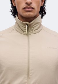 Gros plan d'une personne portant une veste zippée beige avec un col montant et un texte discret sur la poitrine indiquant Peak Performance.