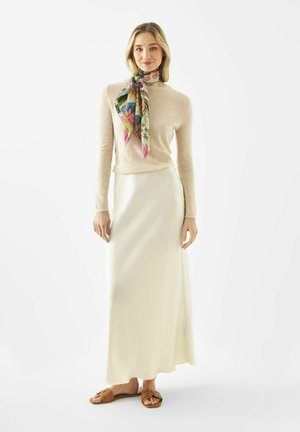 Frau trägt ein beigefarbenes Langarmshirt, einen cremefarbenen Maxirock, einen bunte Blütenschal und braune Sandalen mit offenem Zehenbereich, steht vor weißem Hintergrund.