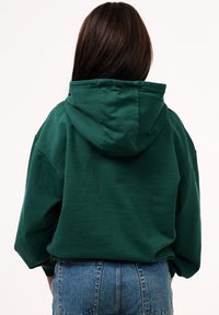 Dunkelgrüner Hoodie mit einer großen, verstellbaren Kapuze. Aus weichem Stoff gefertigt, hat eine lockere Passform und abgesenkte Schulternähte. Getragen mit hochtaillierten Jeans.