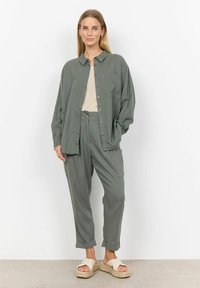 Leichte, olivgrüne Knopfleiste mit einer Brusttasche, kombiniert mit passenden, taillierten Hosen und beigen, strukturierten Sandalen auf einem Betonboden.