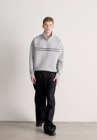 Γκρι φούτερ με φερμουάρ, με μαύρες και ναυτικές ρίγες, με λογότυπο. Φορεμένο με σκούρα μπλε τζιν και μαύρα παπούτσια. Απλό φόντο.