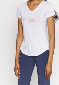 Bílá krátkosrstá tričko s výstřihem do V, které má logo "Columbia Montrail" v růžové a červené barvě, vyrobené z lehké tkaniny s zakřiveným lemem.