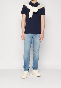 Marinblå polo, ljusblåjeans och vita sneakers. En krämfärgad tröja hängande över axlarna. Enkel design med en klassisk look.