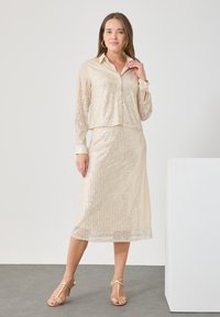 Set de două piese bej, cu mărgele strălucitoare, compus dintr-o bluză scurtă, cu nasturi și mâneci lungi, și o fustă de lungime medie, ambele strălucind cu un model în dungi.