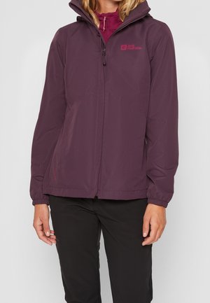 Mujer vistiendo una chaqueta impermeable de color burdeos de Jack Wolfskin con capucha, cerrada con cremallera, combinada con pantalones negros, sobre un fondo liso.