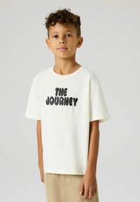 Garçon aux cheveux bouclés portant un t-shirt blanc avec le texte "THE JOURNEY" et un pantalon beige, debout devant un fond clair uni.