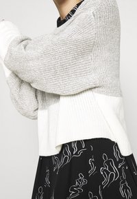 Pull tricoté gris clair et blanc, coupe ample, poignets côtelés et ourlet raccourci, superposé sur une robe noire ornée de motifs en ligne blanche.