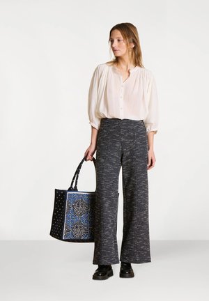 Blouse blanche à boutons avec manches bouffantes, associée à un pantalon large texturé de couleur foncée. Un sac fourre-tout à motifs bleu et noir complète la tenue.