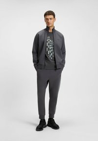 Grå träningsoutfit med en dragkedjans jacka och tapered byxor. Jackan har en hög krage, medan skjortan har ett geometriskt mönster.
