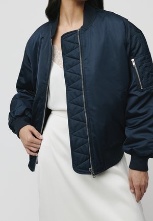 Kobieta nosząca granatową kurtkę bomber na białej sukience z koronkowym wykończeniem, ręce regulujące zamek kurtki.