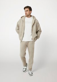 Bežo spalvos zip-up džemperis su kapišonu ir derančios poilsinės kelnės, pagaminti iš minkštos medžiagos, su logotipu ant krūtinės ir šono, įkombinuoti su pilkos ir baltos spalvos sportbačiais.