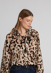 Blusa de estampado de leopardo con textura plisada, cuello recogido y lazo. Colores beige claro y marrón oscuro, con mangas largas y un corte holgado.