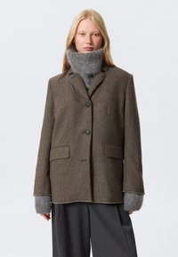 Bruine herringbone blazer met twee voorzakken, knoopsluiting en grijze gebreide manchetten. De kraag heeft een licht rafelige rand.