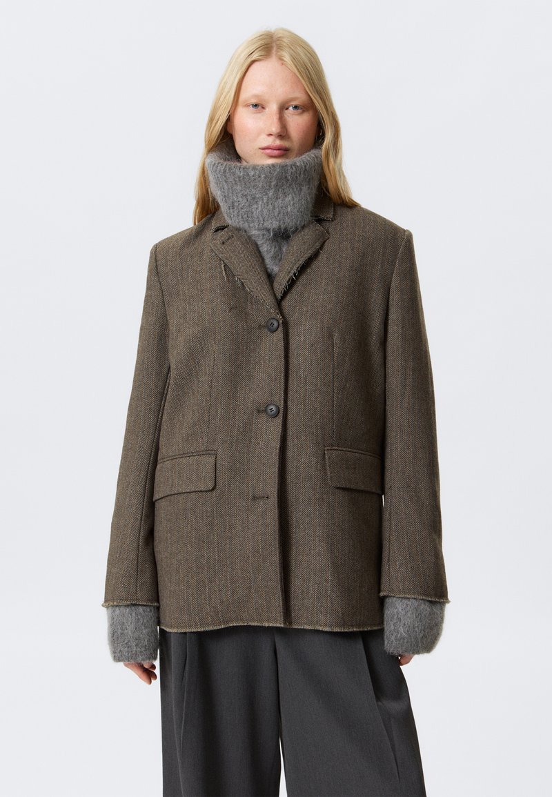 Bruine herringbone blazer met twee voorzakken, knoopsluiting en grijze gebreide manchetten. De kraag heeft een licht rafelige rand.
