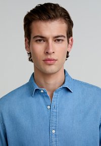 Camasa din denim bleu deschis, cu guler cu nasturi și nasturi albi. Prezintă un logo brodat pe piept și o textură subtilă, cu o țesătură netedă.