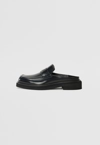 Zapato negro de cuero sin cordones con puntera redondeada, un panel delantero elevado y suela de goma gruesa. Textura suave con un detalle mínimo.