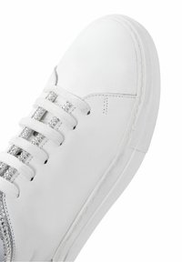 Valkoinen nahkakengän malli, jossa on tasaiset nauhat ja kimaltava hopeinen yksityiskohta sivussa. Rikkaita kumipohja pyöreällä varvasyhteydellä.