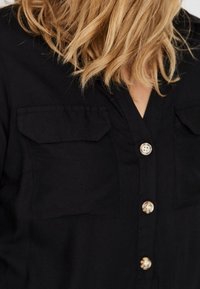 Camisa negra de botones con dos bolsillos en el pecho, un escote en V y una textura de tela suave. Detalles notables incluyen botones plateados.